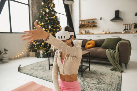Foto de Una encantadora joven es vista estirándose junto a un árbol de Navidad mientras usa gafas de realidad virtual. Foto de alta calidad - Imagen libre de derechos