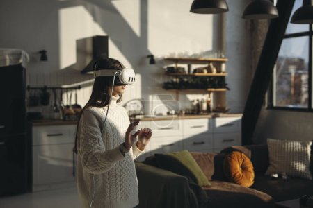 Téléchargez les photos : Dans le confort de son appartement, une superbe jeune femme joue à un jeu en ligne avec un casque VR. Photo de haute qualité - en image libre de droit