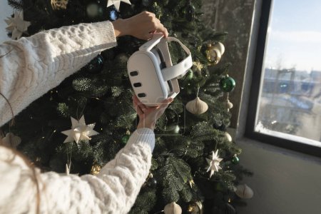 Photo for Against the setting of a Christmas tree, a girl holds a virtual reality headset in her hands. High quality photo - Royalty Free Image