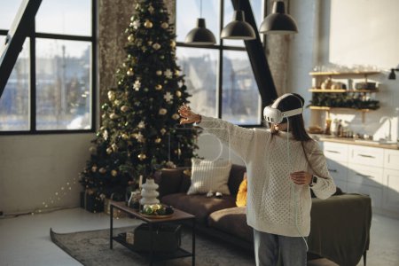 Foto de Absorto en un juego en línea, una encantadora joven lleva un auricular de realidad virtual en su apartamento. Foto de alta calidad - Imagen libre de derechos