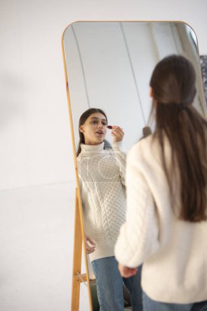 Foto de Con alegría navideña alrededor, una joven impresionante trabaja en su maquillaje frente al espejo. Foto de alta calidad - Imagen libre de derechos