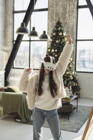 Téléchargez les photos : Embrassant l'esprit festif, une belle jeune femme utilise un casque VR près d'un arbre de Noël. Photo de haute qualité - en image libre de droit