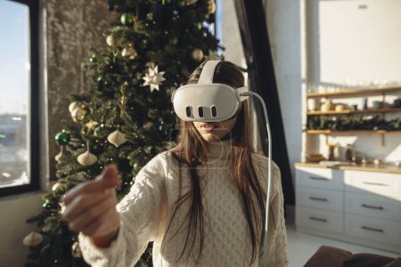 Téléchargez les photos : Portrait d'une jolie fille dans un casque de réalité virtuelle avec un arbre de Noël en arrière-plan. Photo de haute qualité - en image libre de droit