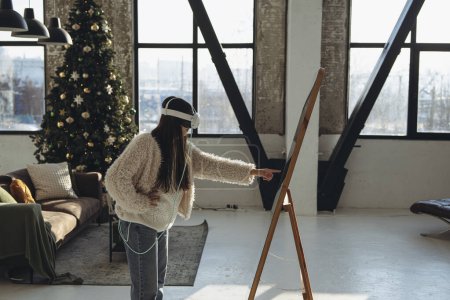 Téléchargez les photos : Au milieu de l'aura de Noël, une jeune femme en vêtements légers et un casque de réalité virtuelle se tient devant le miroir. Photo de haute qualité - en image libre de droit
