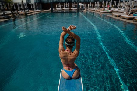 Téléchargez les photos : Une charmante femme en bikini est assise au bord de la piscine, le dos contre le bord. Photo de haute qualité - en image libre de droit