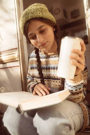 Téléchargez les photos : Une fille branchée et inspirée des hippies savoure une boisson chaude sur la terrasse de la maison, mettant en valeur son style. Photo de haute qualité - en image libre de droit