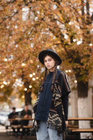 Foto de Una mujer encantadora, de estilo boho, lleva un sombrero negro mientras pasea por las calles de la ciudad de otoño. Foto de alta calidad - Imagen libre de derechos