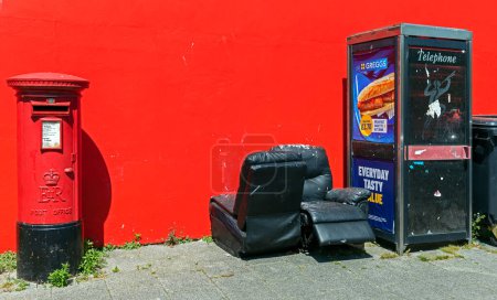 Téléchargez les photos : Une scène de rue avec une cabine à piliers, une cabine téléphonique et des meubles jetés contre un mur rouge. - en image libre de droit