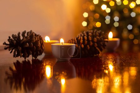 Foto de Decoración de Navidad. Algunas velas encendidas en una superficie reflectante, dos conos de pino y un detalle desenfocado del árbol de Navidad en el fondo. - Imagen libre de derechos