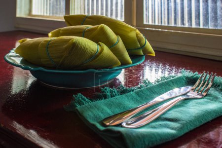 Téléchargez les photos : 3 pamonhas recouverts d'écorces de maïs, à l'intérieur d'une assiette, devant une fenêtre et sur une surface brillante. - en image libre de droit