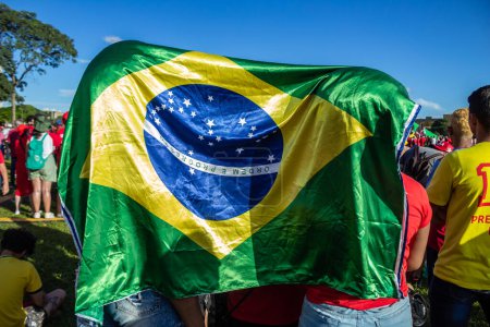 Foto de Two people using a Brazilian flag to protect themselves from the sun. Photo taken at the inauguration event of the new president of Brazil. - Imagen libre de derechos