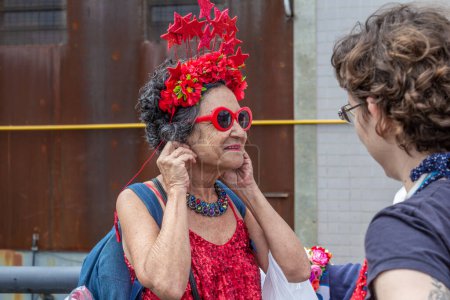 Foto de Una anciana, sonriendo, ajustando pendientes en las orejas y con una tiara de flores rojas en la cabeza. Foto tomada durante el acto de inauguración del Presidente. - Imagen libre de derechos