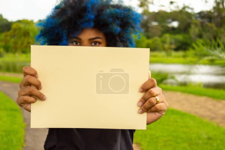 Photo for A young woman, with hair dyed blue, her face hidden behind a blank poster, with a landscape in the background. - Royalty Free Image