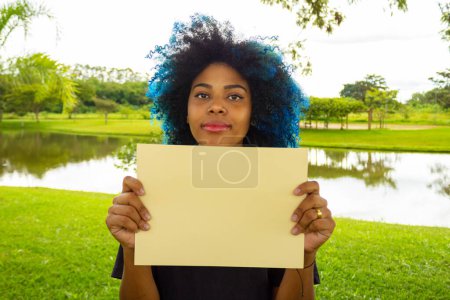 Photo for A young woman, with dyed blue hair, holding a blank sign, with a landscape in the background. - Royalty Free Image