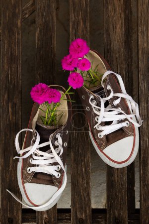 Foto de Un par de zapatillas negras viejas y descoloridas, con flores sobresaliendo de ellas, sobre una superficie de clavijas de madera. - Imagen libre de derechos