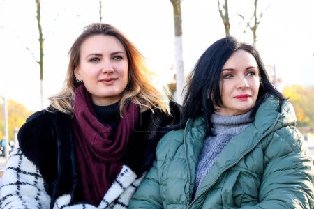 Téléchargez les photos : Portrait de deux femmes assises dehors sur un banc en manteaux - en image libre de droit