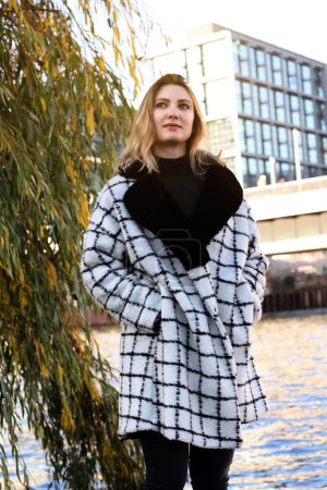 Photo for Young woman in a coat walking along a river in the city - Royalty Free Image