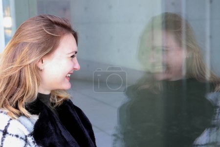 Foto de Joven mujer de pie fuera y mirando su reflejo de risa en la ventana - Imagen libre de derechos