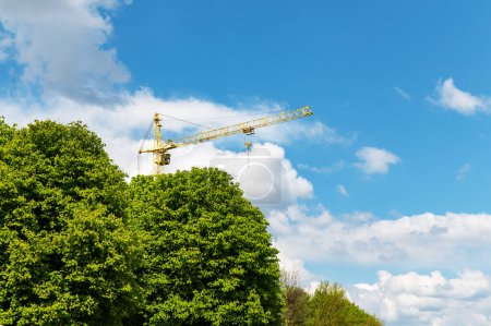 Grúa de construcción industrial detrás de los árboles contra el cielo con nubes blancas. Grúa de construcción en la distancia se puede ver desde detrás de las copas de los árboles de verano