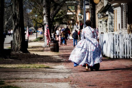 Photo for A woman, in period dress, walks behind tourists on East Duke of Gloucester Street in Colonial Williamsburg. - Royalty Free Image