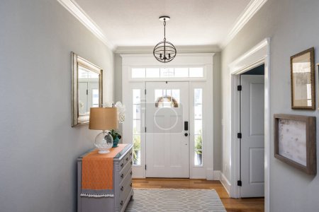 Téléchargez les photos : An open large and wide interior front door hallway foyer with transom, hanging light fixture, coastal colors and entry way table and wood floors. - en image libre de droit