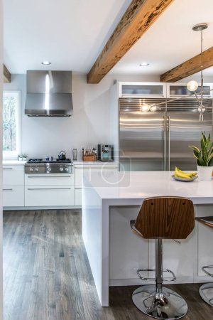 Une grande cuisine moderne de luxe avec murs blancs et poutres en bois et une grande île de quartzite de quartz avec des appareils haut de gamme.