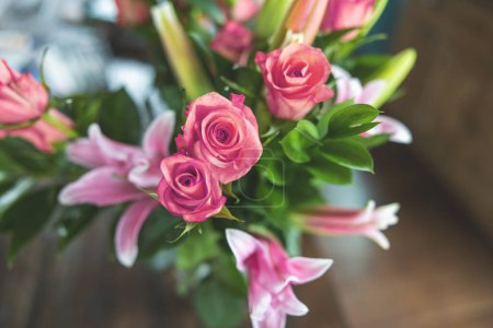 Téléchargez les photos : Un beau bouquet de roses et de lys roses et corail dans un pichet de vase clair sur une table de salle à manger en bois sombre avec lumière naturelle si les fenêtres. - en image libre de droit