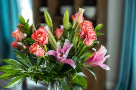 Téléchargez les photos : Un beau bouquet de roses roses et de corail et de lis dans un pichet de vase clair sur une table de salle à manger en bois sombre avec de la lumière naturelle venant par les fenêtres. - en image libre de droit
