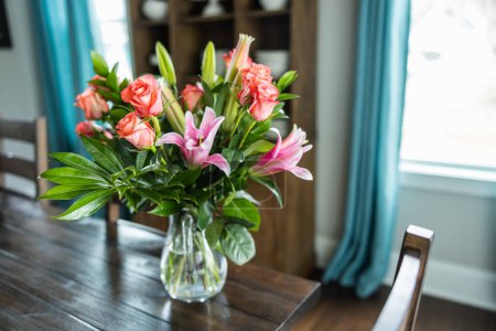 Téléchargez les photos : Un beau bouquet de roses roses et de corail et de lis dans un pichet de vase clair sur une table de salle à manger en bois sombre avec de la lumière naturelle venant par les fenêtres. - en image libre de droit