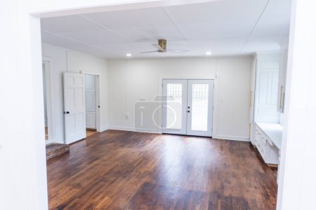 Empty white living room or den of a newly renovated and painted house with dark hardwood floors and double window doors out to the patio.