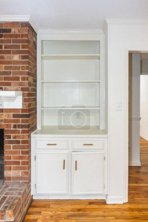 An empty white built in bookcase in a living room next to a red brick fireplace.