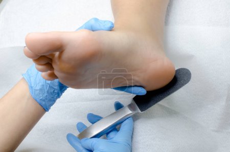 Téléchargez les photos : Pédicure en gants bleus pratiquant une pédicure médicale professionnelle dans un salon de beauté. Traitement des pieds avec râpe spéciale pour frotter les talons de près - en image libre de droit