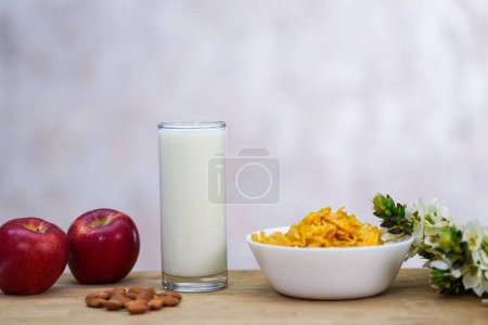 Téléchargez les photos : Cornflakes dans un bol blanc, un verre de lait, graines d'amandes, pains de blé entier, pomme rouge et fleurs blanches sur la table en bois. - en image libre de droit
