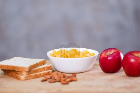 Téléchargez les photos : Cornflakes dans un bol blanc avec lait, graines d'amandes, pains de blé entier et pomme rouge sur la table en bois. - en image libre de droit