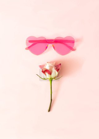 Pink heart shaped sunglasses and a white and magenta rose on pastel pink background. Vertical design for wallpaper. Flat lay