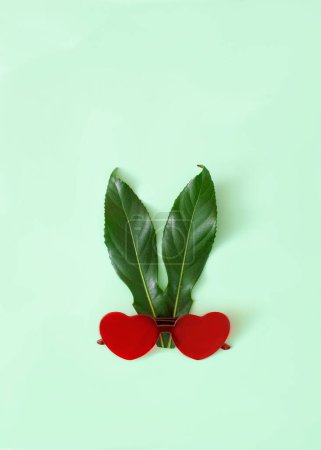Foto de Gafas de sol rojas en forma de corazones y hojas verdes como orejas sobre fondo verde pastel. Concepto surrealista mínimo para tarjeta o banner de San Valentín. Diseño para anuncio de celebración de Pascua. - Imagen libre de derechos