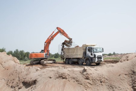 Foto de Excavadora de orugas de color naranja grande y un camión volquete de construcción de pie lado a lado mientras se trabaja en un sitio de construcción en un pozo de arena, el suelo está siendo cargado y transportado por un camión - Imagen libre de derechos