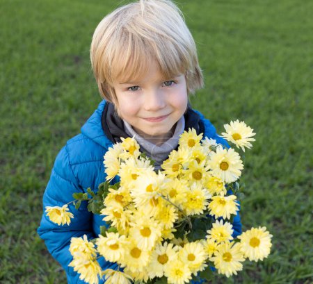 Téléchargez les photos : Portrait de garçon souriant en veste bleue avec bouquet de chrysanthèmes jaunes. Fête des mères, fleurs de fils. humeur festive, gratitude. Gloire à l'Ukraine. Arrêtez la guerre. Soutenir l'Ukraine, paix aux Ukrainiens - en image libre de droit