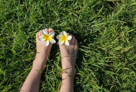 Téléchargez les photos : Pieds nus de l'enfant reposent sur une pelouse verte, deux fleurs plumeria entre les orteils. joie, atmosphère positive joyeuse, enfance heureuse, repos. Bonjour l'été. l'énergie de la nature. Jour de la Terre - en image libre de droit