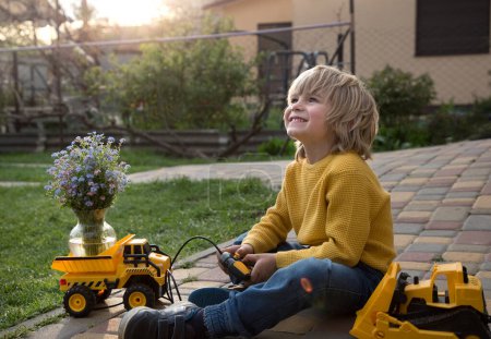 Téléchargez les photos : Cute smiling boy 6 years old plays with a toy dump truck and a bulldozer. driving a bouquet of flowers for mom on mother's day. builder's holiday, motorist's day, humorous delivery advertising - en image libre de droit