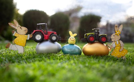 Foto de Two models of toy tractors stand on painted multi-colored eggs that lie on the grass. concept of commercial easter greetings for farms, agribusiness. postcard. copy space - Imagen libre de derechos