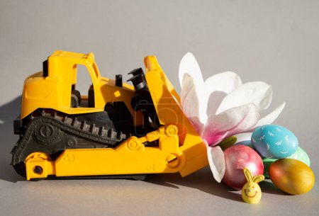 Foto de Toy orange bulldozer, colorful Easter eggs, magnolia flower. The concept of congratulations on the Easter holiday for business, construction companies - Imagen libre de derechos