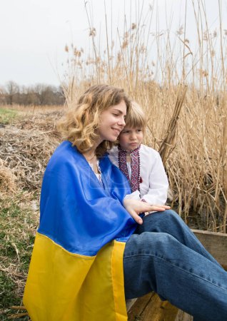 Téléchargez les photos : Petit garçon et une femme enveloppés dans le drapeau ukrainien s'embrassent, à la recherche de soutien. Famille, réfugiés, unité, soutien, patriotisme, nostalgie. Arrêtez la guerre en Ukraine. Fierté d'être ukrainien - en image libre de droit