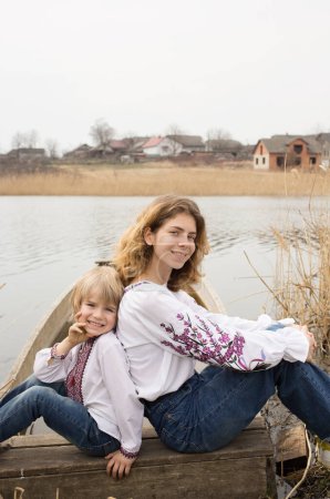 Téléchargez les photos : Heureux petit garçon et jeune belle femme en vêtements nationaux brodés assis à côté de l'autre sur la rive du lac. Famille, réfugiés, unité, soutien, liberté, indépendance. Ukrainiens contre la guerre - en image libre de droit