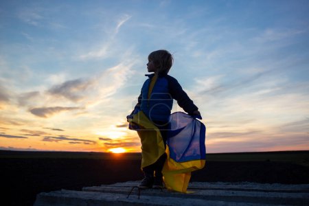 Silhouette eines traurigen Jungen mit ukrainischer Flagge vor dem Hintergrund des strahlend schönen Sonnenuntergangs. Kinder gegen den Krieg. Stoppt die russische Aggression. Das Konzept des Patriotismus und der Vaterlandsliebe