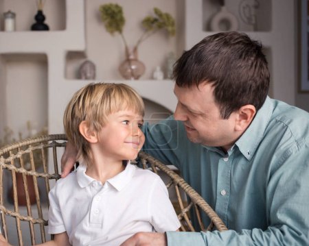 Photo for Happy father is sitting next to his preschool son, smiling, looking into each other's eyes. dad and little boy enjoy the weekend at home, relaxing, chatting with family. father's day - Royalty Free Image