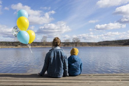 Téléchargez les photos : Méconnaissable petit garçon et femme assis avec le dos sur le rivage d'un lac avec des ballons bleus et jaunes. Famille, réfugiés, unité, soutien, patriotisme, nostalgie. stand avec l'Ukraine - en image libre de droit