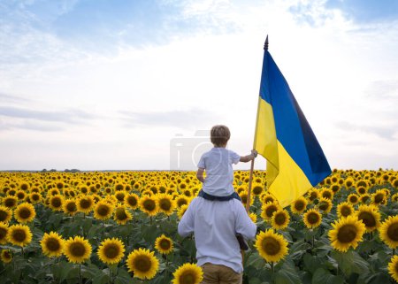 Photo for Unrecognizable boy sitting on shoulders of man stands with large flag of Ukraine among blooming field of yellow sunflowers. Education of patriotism. Pride, faith in victory. stop war. Independence Day - Royalty Free Image