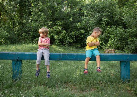 Photo for Two brothers or boys friends in casual clothes are sitting on bench offendedly turned away in different directions, sad and gloomy. Offended, dissatisfied, do not want to agree with each other. - Royalty Free Image