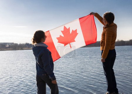 Foto de Dos mujeres sostienen la bandera canadiense, en un día soleado sobre el fondo del cielo y el río. Viajes, inmigración. Día de Canadá. Orgullo, libertad, símbolo nacional - Imagen libre de derechos